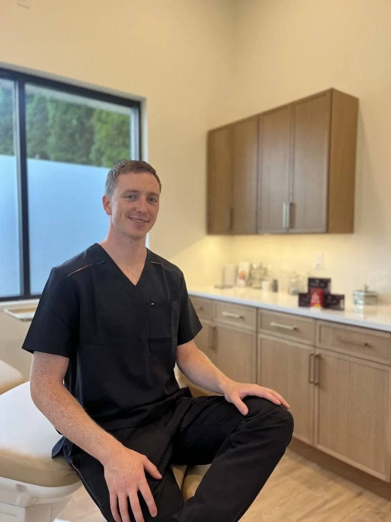 A young man in black medical scrubs sitting on a beige examination table in a room with wooden cabinets, a large window, and a countertop with medical supplies.