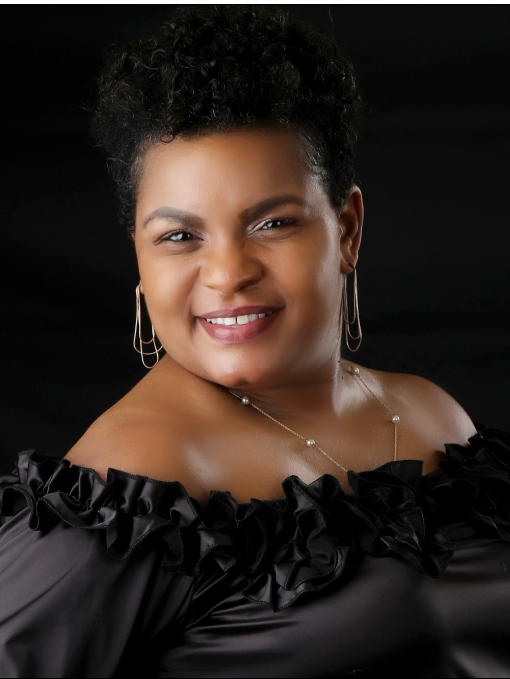 A woman with curly black hair smiling at the camera, wearing a black off-shoulder dress with ruffles and gold jewelry against a black background.