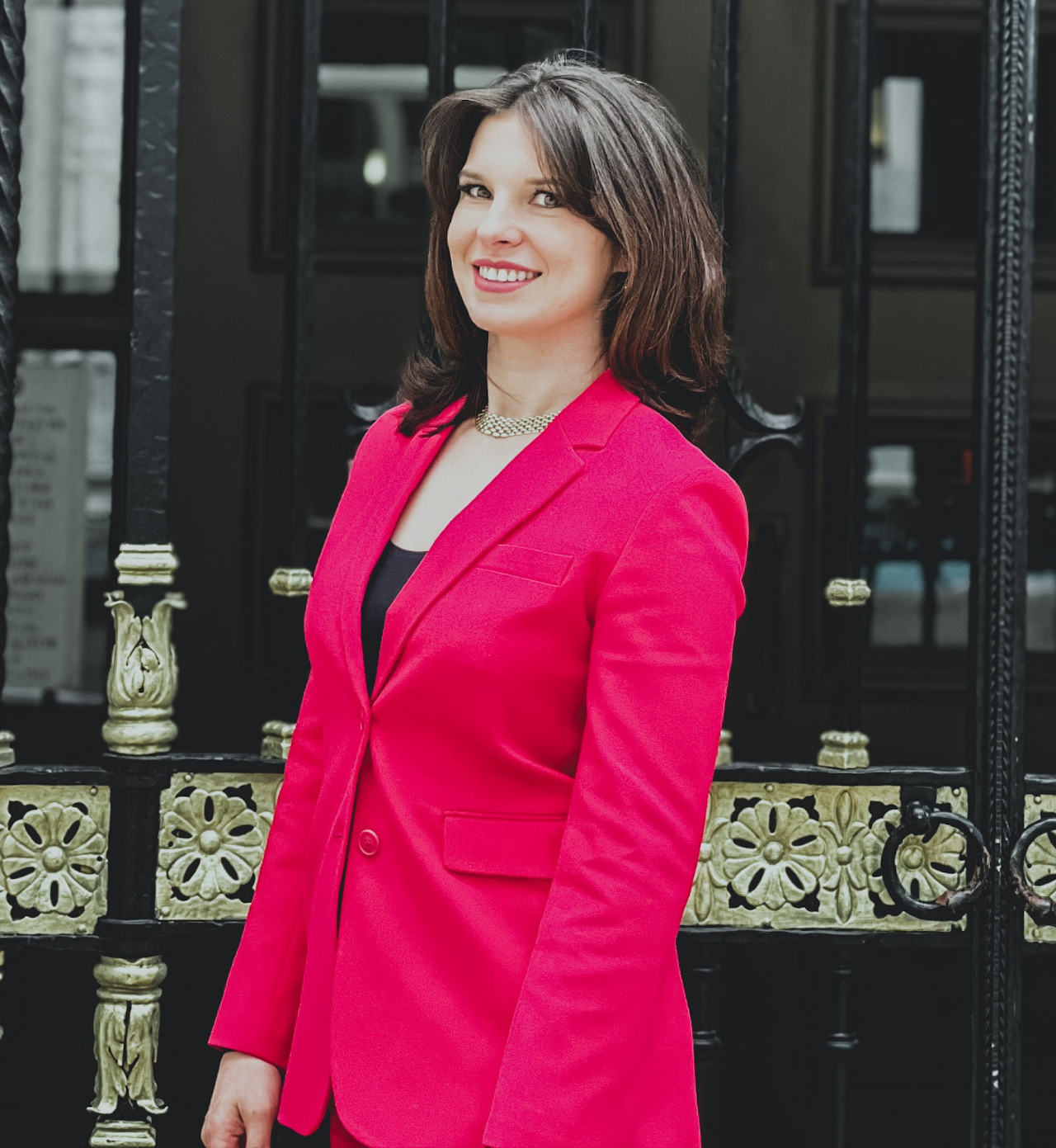 A woman in a bright pink blazer and black top standing outdoors in front of a decorative black and gold gate, smiling at the camera.