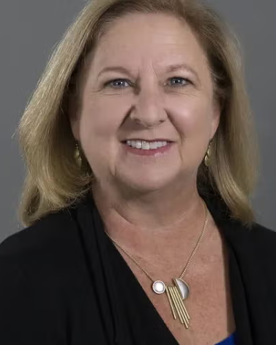 A smiling middle-aged woman with blonde hair, wearing a black top, gold earrings, and a necklace with circular and fringe design, posed against a gray background.