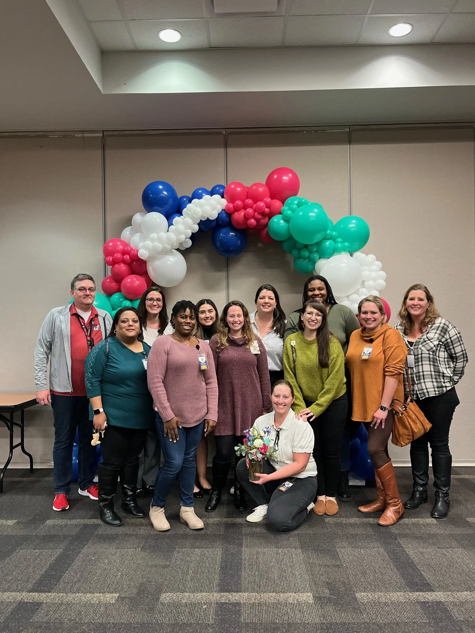 A group of diverse women and one man posing together indoors in front of colorful balloon arch, some wearing badges and one woman holding a bouquet of flowers.