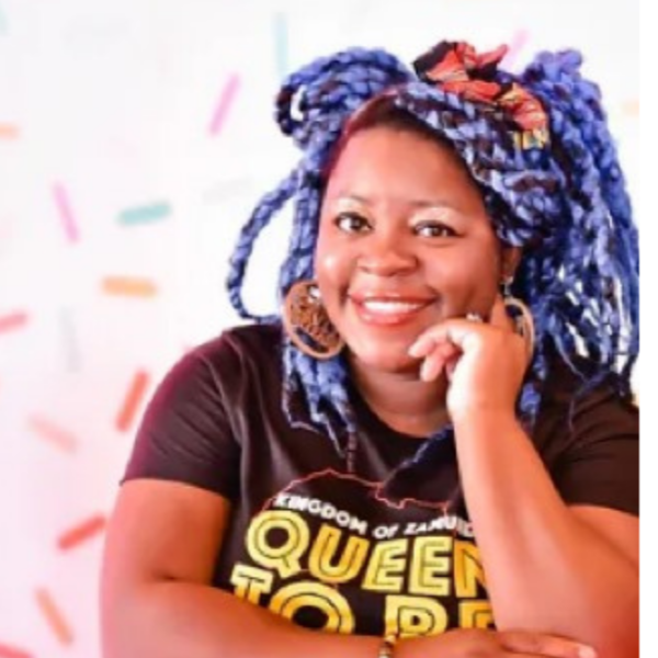 Woman with blue and purple dreadlocks, wearing large earrings, smiling, resting her chin on her hand, sitting against a colorful background.