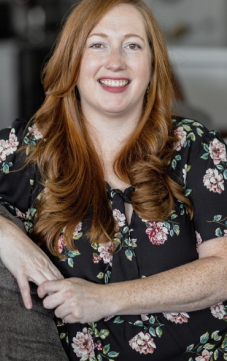 Woman with long red hair smiling, wearing a black floral top, sitting on a gray couch in a modern living room.