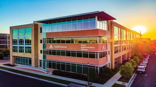 Modern office building with glass windows and a sign reading 'Ascension Texas Administrative Offices' during sunset.