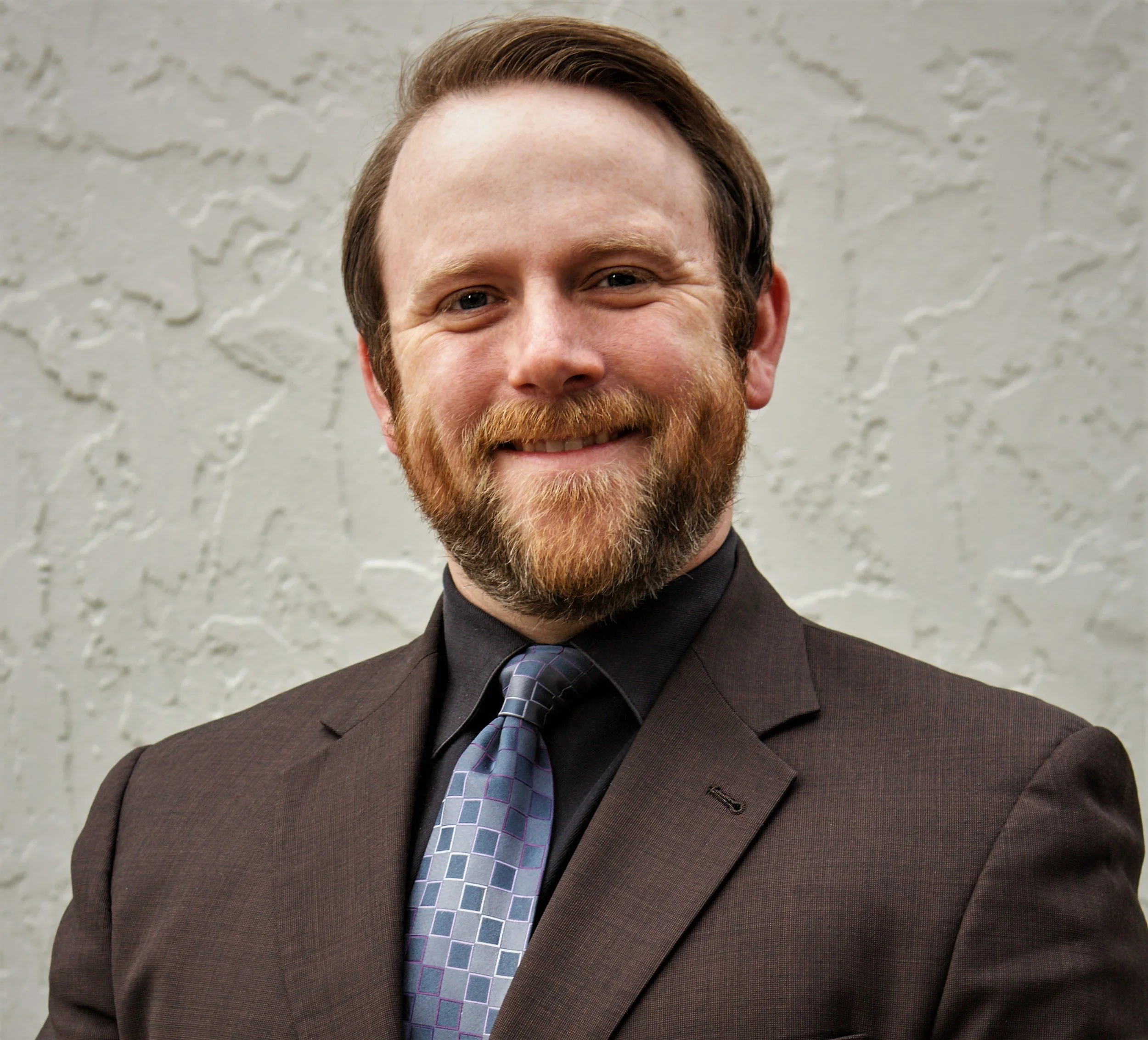 Close-up of a man in a dark suit and patterned tie, smiling, with a textured light-colored wall in the background.