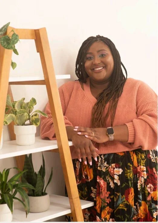 Smiling woman with long braids wearing a peach sweater and a floral skirt, standing next to a white shelf with potted plants