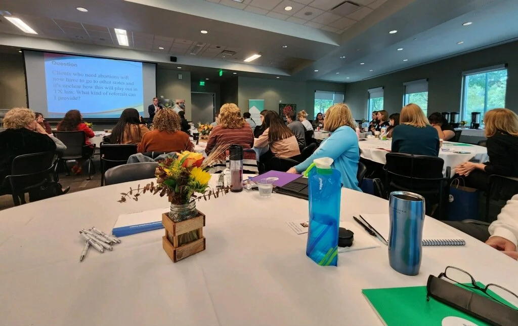 A conference room filled with attendees seated at round tables, listening to a speaker on stage. The room has large windows, and a projection screen displays a presentation.