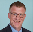 A smiling man in a suit with glasses, short red hair, and a blue shirt against a plain background.