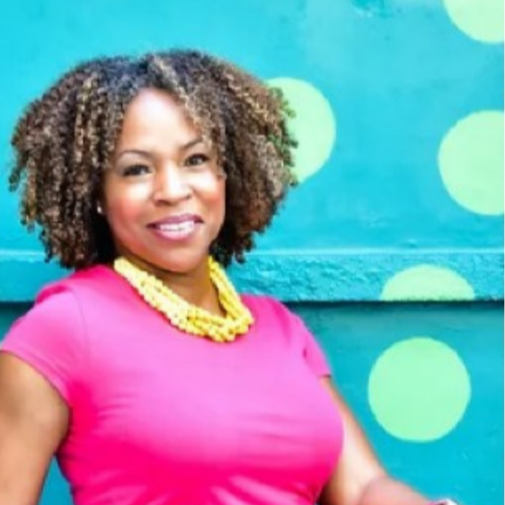 Woman with curly hair wearing a pink top and yellow necklace, standing in front of a teal wall with large yellow polka dots.