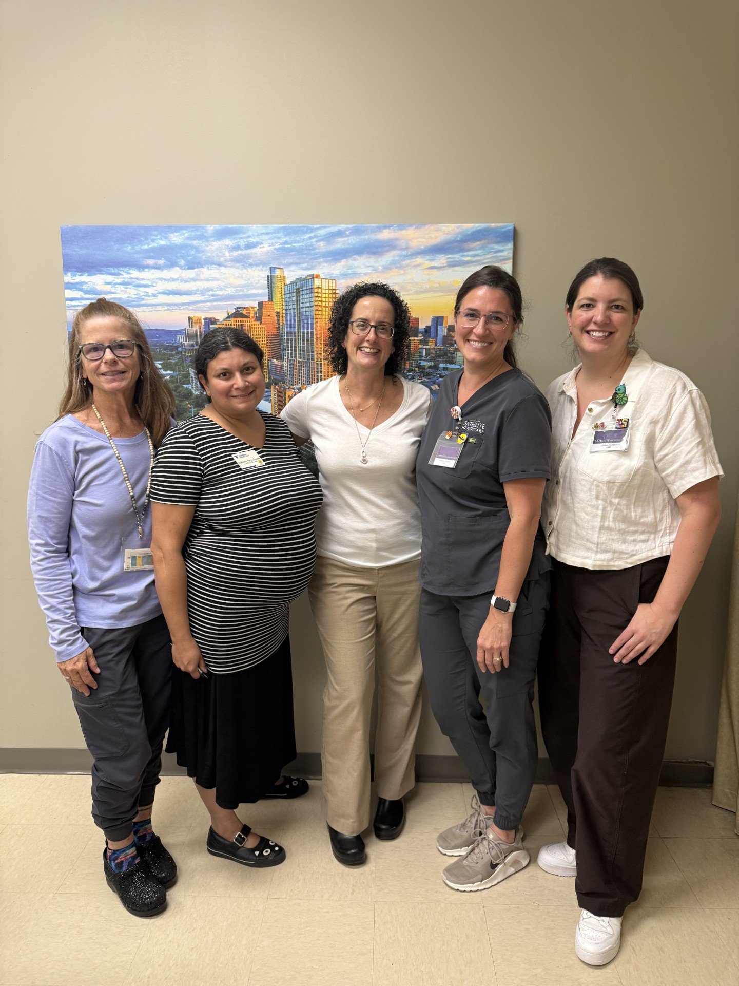 Five women standing together in a hospital hallway, smiling, with a cityscape picture on the wall behind them.