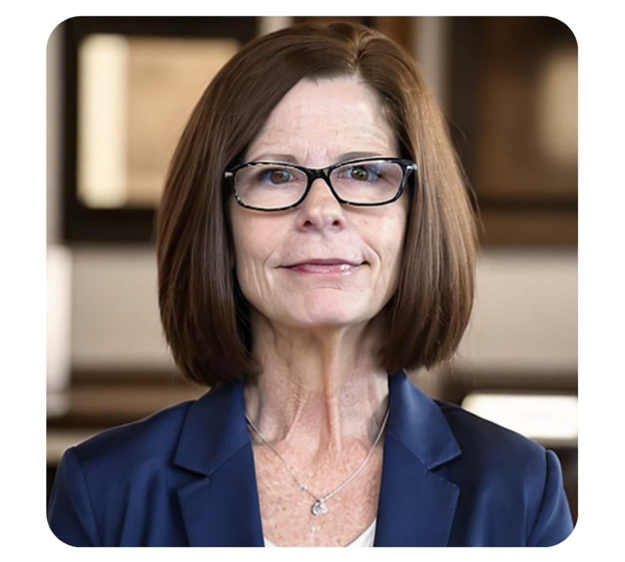 A middle-aged woman with shoulder-length brown hair, wearing glasses and a navy blazer, smiling slightly in an indoor setting.