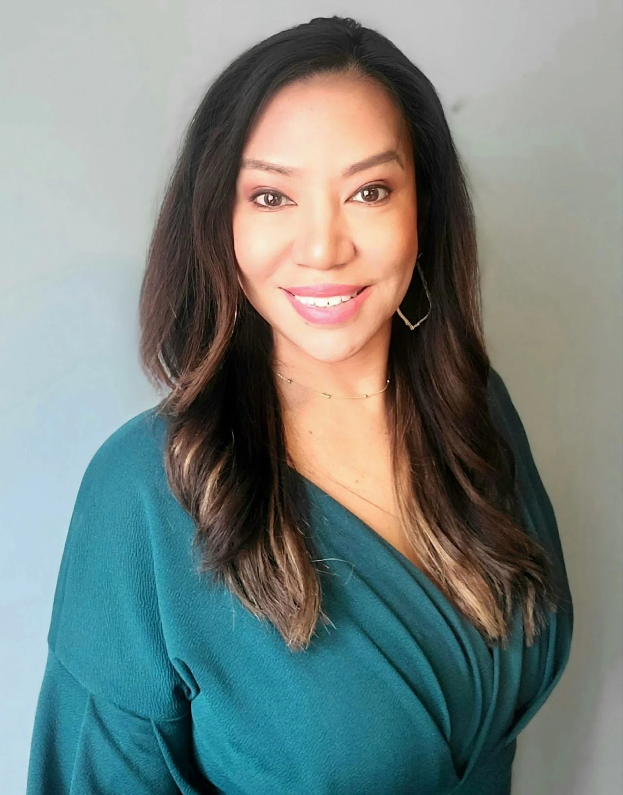 A woman with long dark hair and light skin smiling, wearing a teal blouse, gold jewelry, standing against a plain light gray background.