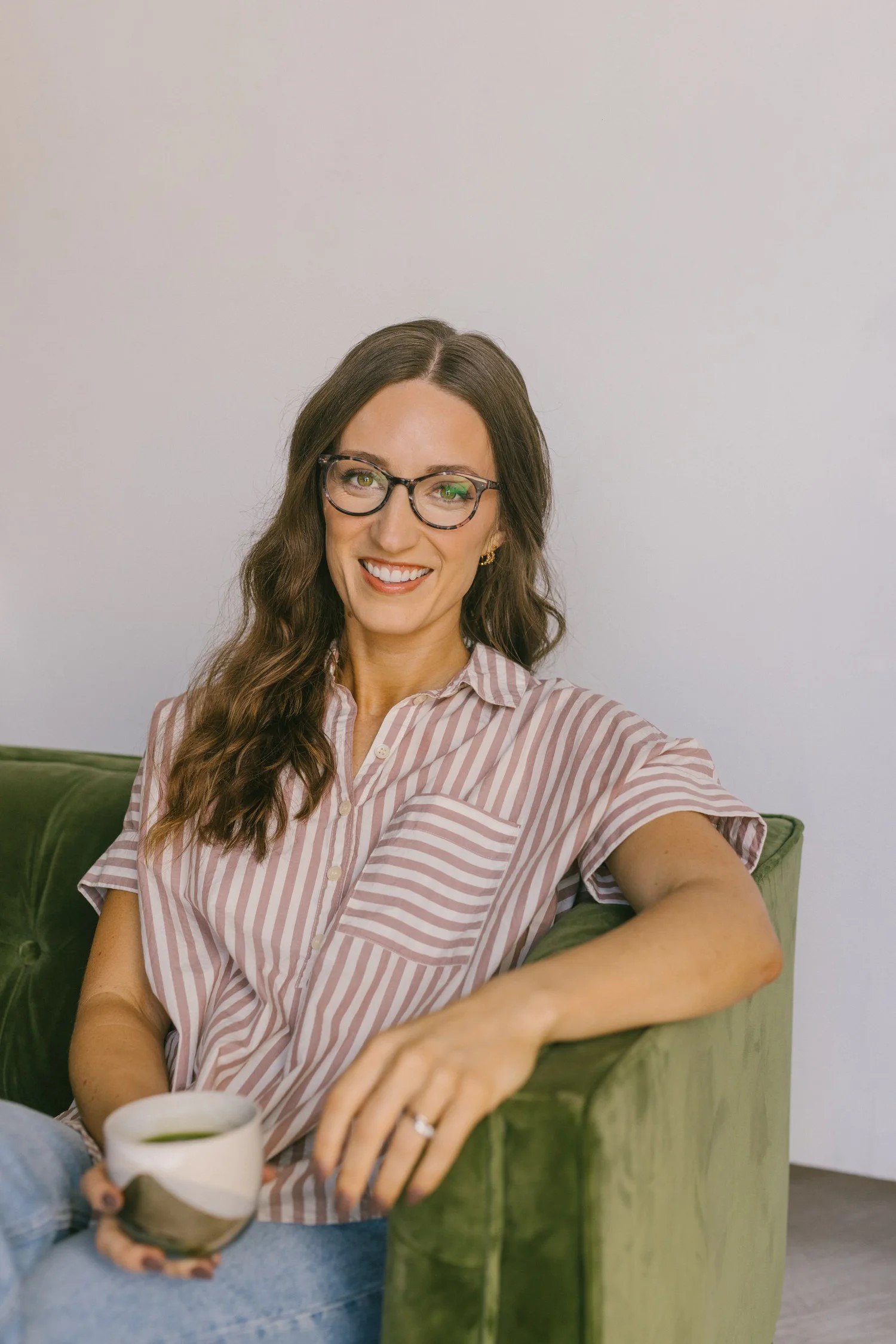 Woman with brown hair, glasses, and a striped pink and white shirt, sitting on a green couch, smiling, holding a white mug with a hot beverage, in a room with white walls.