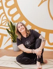 woman sitting on a mat in a yoga studio with a large wall clock design and a potted plant in the background, smiling