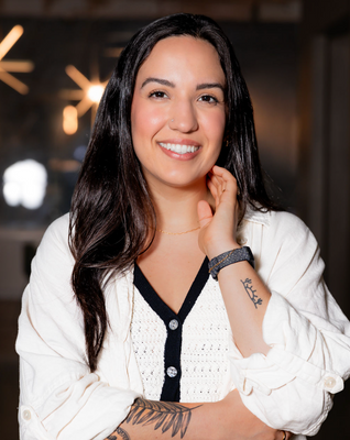 Portrait of a woman with long dark hair, smiling, wearing a white cardigan with navy trim, in an indoor setting.