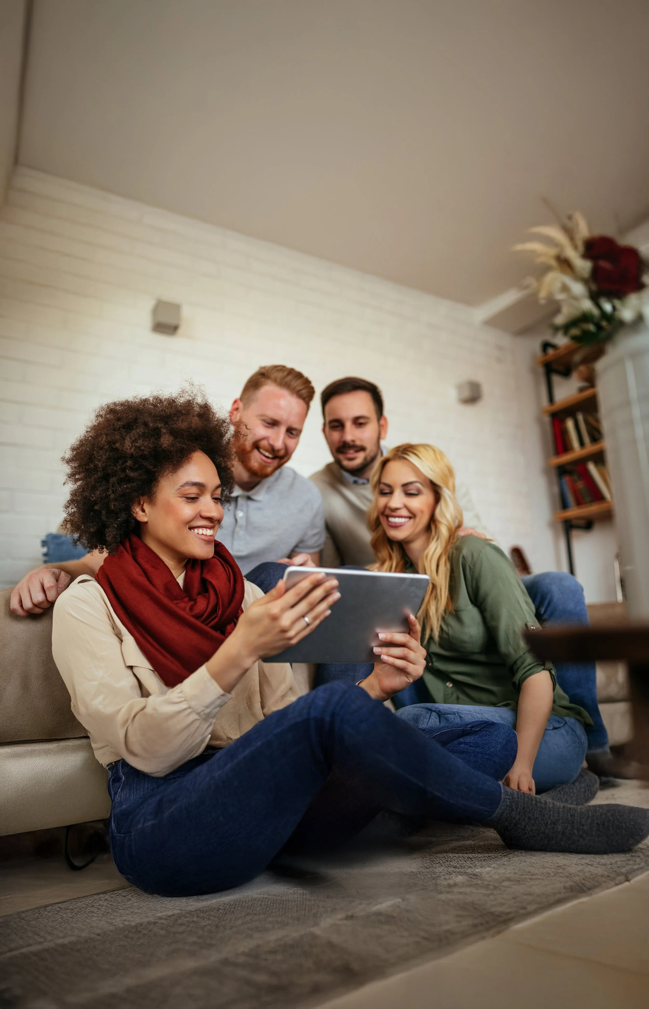Group of five young adults sitting on the floor in a cozy living room, looking at something on a tablet and smiling.