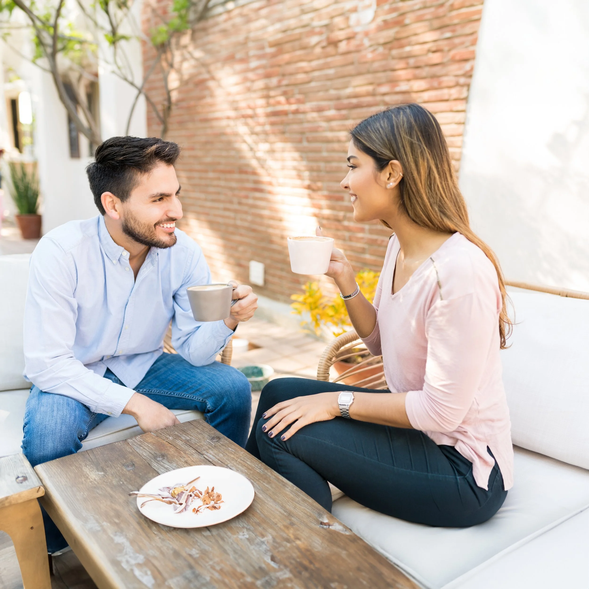 A man and woman sitting outside at a wooden table, smiling and talking while holding cups. There is a partially eaten pastry on a plate on the table. They are enjoying a casual conversation in a cozy outdoor cafe setting.
