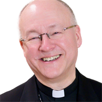 A smiling man dressed as a priest or clergy member wearing glasses and a black shirt with a clerical collar.