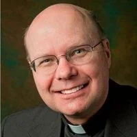 Close-up portrait of a smiling middle-aged man with glasses, light skin, and short hair, wearing a dark suit and clerical collar.