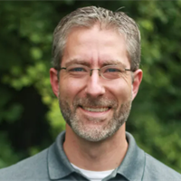 A smiling man with glasses and gray hair outdoors near greenery.