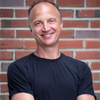 A smiling man in a black t-shirt standing outdoors in front of a brick wall.