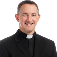 Portrait of a smiling man dressed as a priest in a black suit and clerical collar.