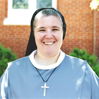 A woman in a light blue uniform and a cross necklace smiling outdoors with a brick building and greenery in the background.