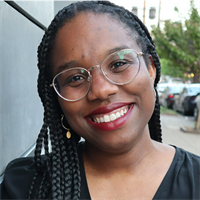 Close-up of a smiling woman wearing glasses, with braided hair and gold earrings outdoors.