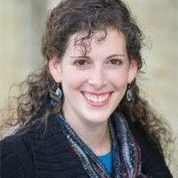 A woman with curly hair smiling, wearing earrings and a dark top, outdoors with greenery in the background.