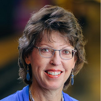 A woman with glasses, curly hair, and earrings smiling at the camera.