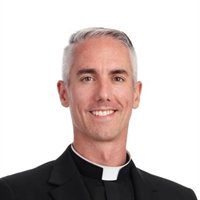 Portrait of a smiling middle-aged man with short gray hair, wearing a black clerical shirt with a white collar.