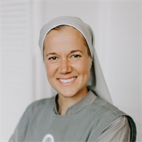A smiling woman dressed as a nurse, wearing a light gray uniform and a white head covering, standing indoors against a neutral background.