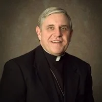 Photograph of a smiling middle-aged man dressed as a Catholic priest, wearing a black suit with a clerical collar, against a neutral background.
