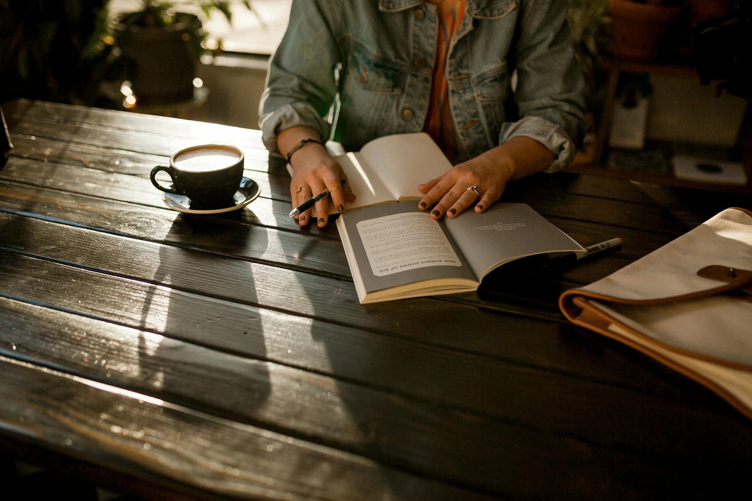 Person sitting at a wooden table with a cup of coffee and open books, studying or reading, in a cozy setting with warm lighting.