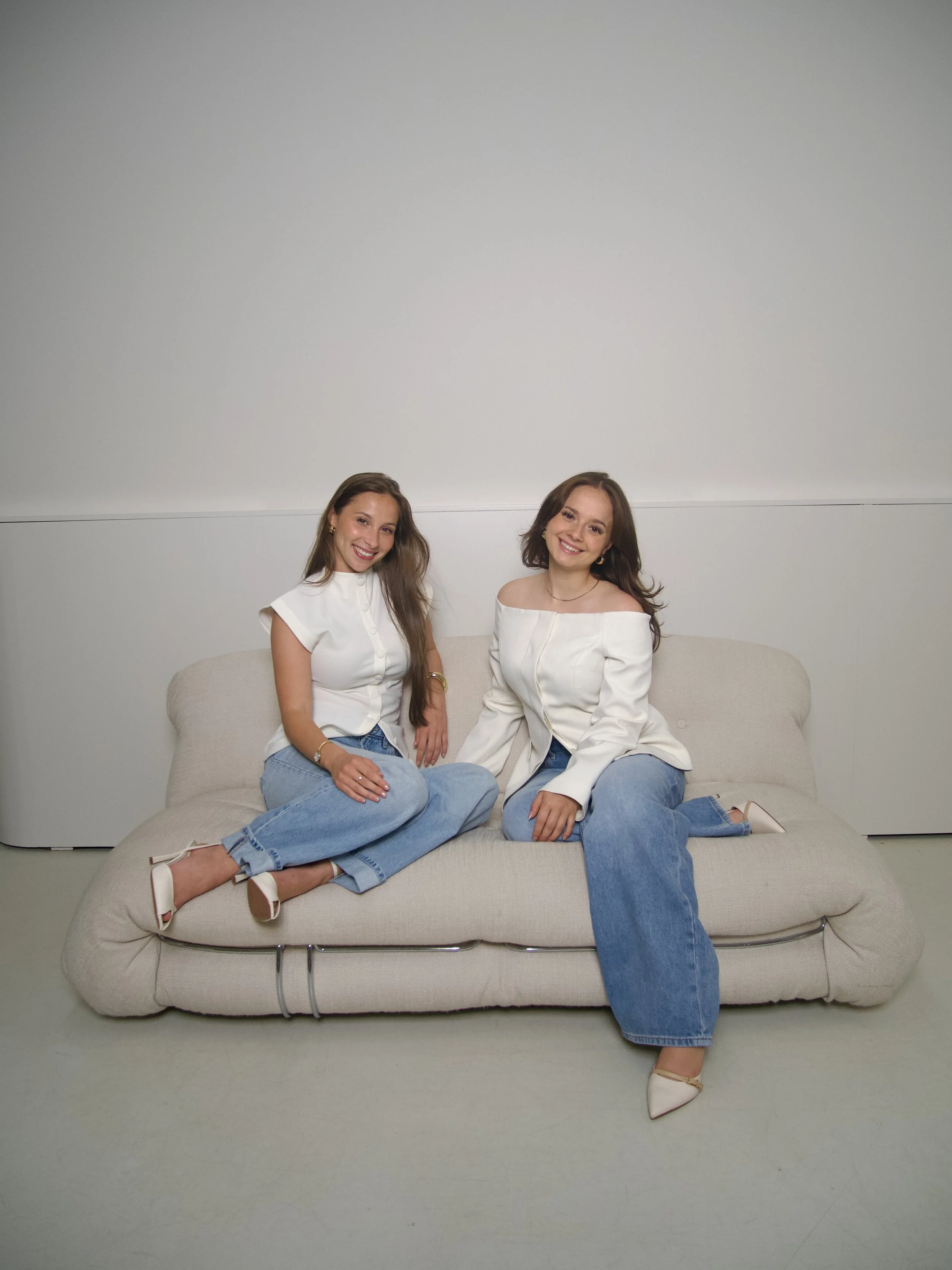 Zwei junge Frauen sitzen entspannt auf einem Sofa, lächeln und schauen in die Kamera. Sie tragen weiße Oberteile und blaue Jeans.