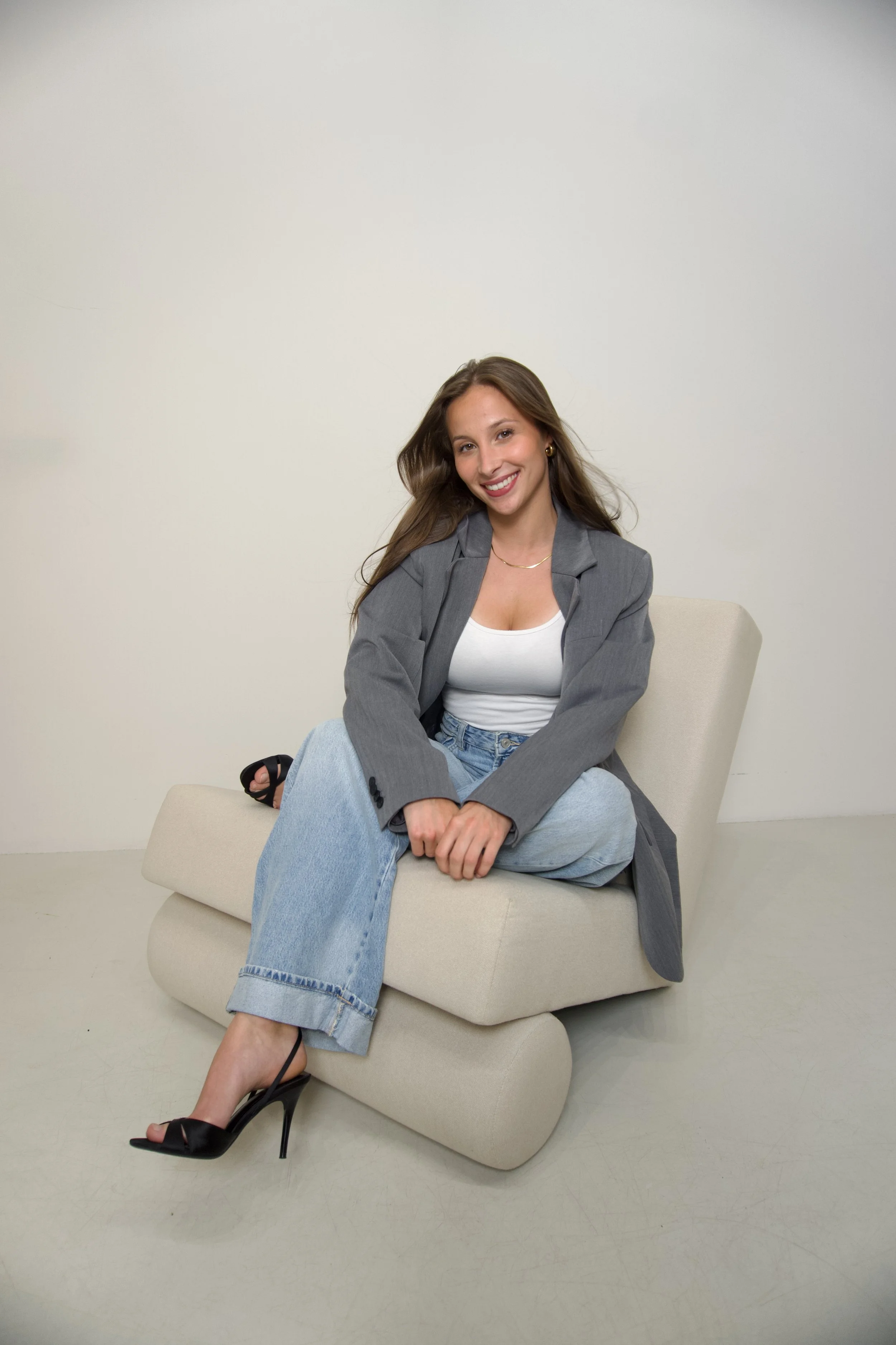 Junge Frau mit langen braunen Haaren, grauem Blazer, weißem Top, weiten Jeans und schwarzen High Heels sitzt auf einem modernen beigen Sessel vor weißem Hintergrund, lächelt freundlich.