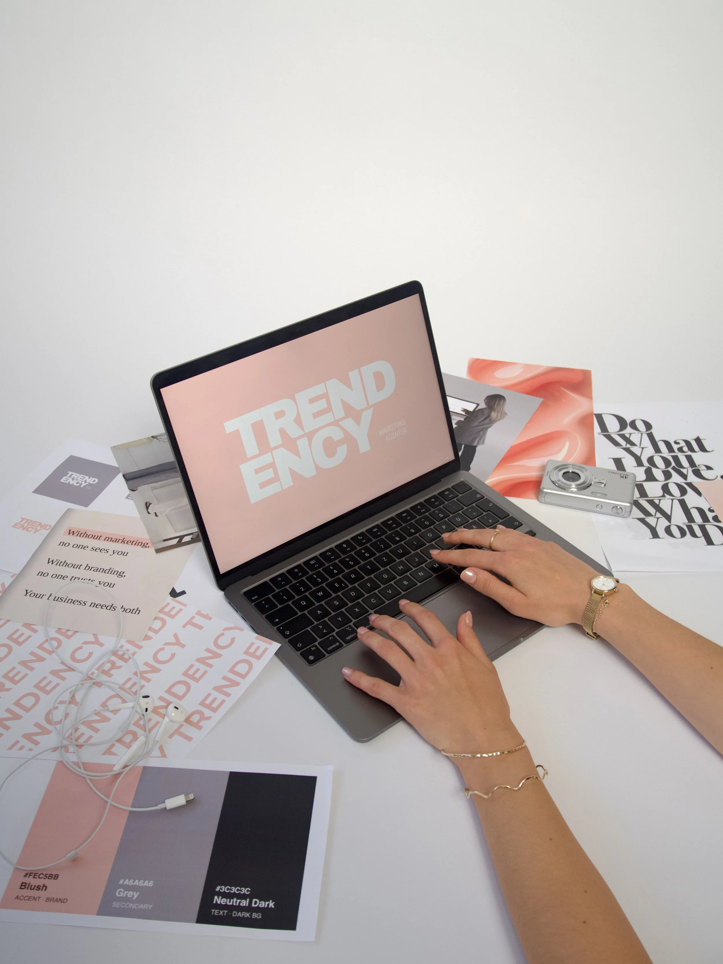 Ein Laptop mit der Anzeige 'Trend Agency', umgeben von gedruckten Farbmustern und Texten, sowie einer Kamera und Kopfhörern auf einem weißen Tisch.