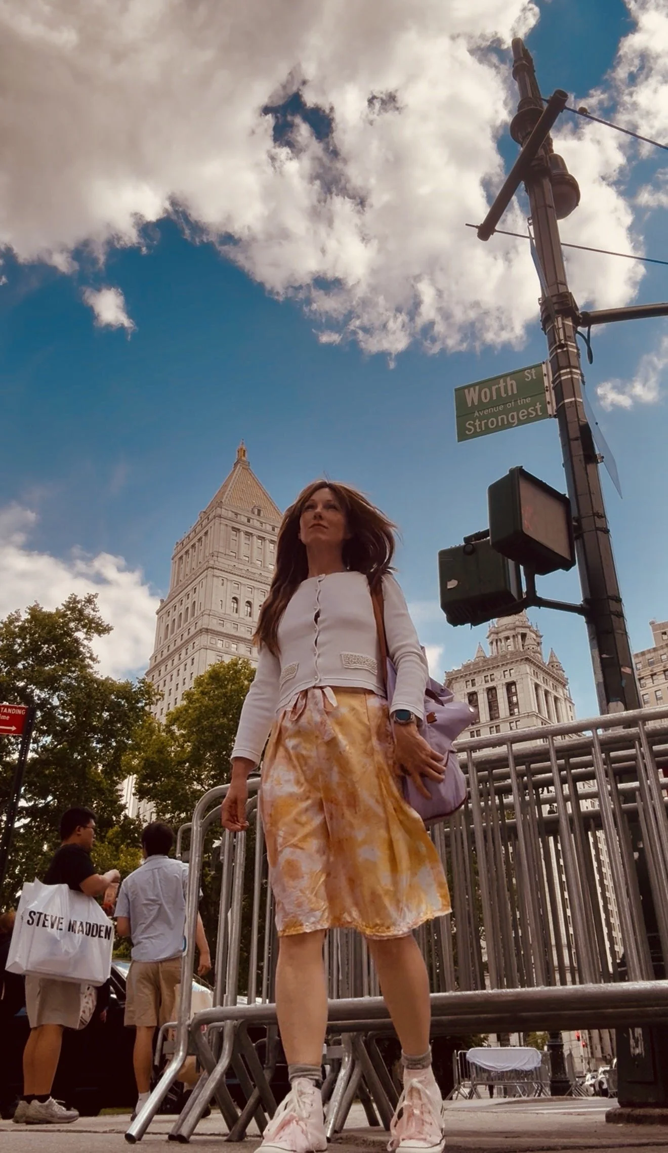 Woman with long brown hair standing on metal barricades in front of city buildings, including the Flatiron Building, under a partly cloudy sky, wearing a white top, orange and yellow tie-dye skirt, and pink chunky sneakers, with a street sign that reads Worth Street.