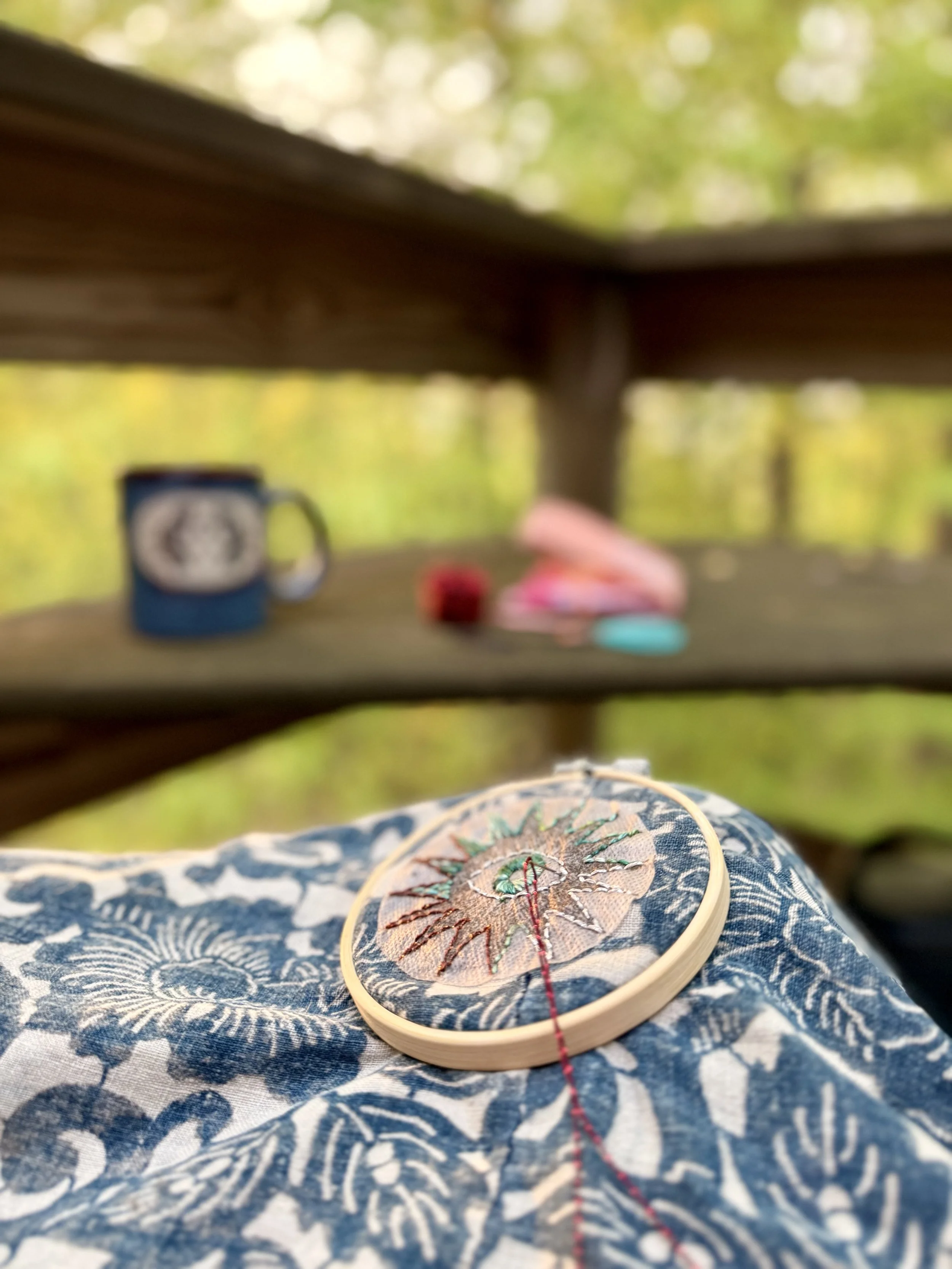 Close-up of embroidery on fabric with floral design, placed on a patterned fabric surface, with outdoor greenery in the background.