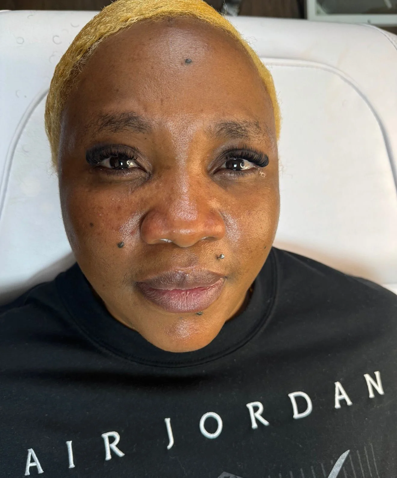A woman with short blonde hair and dark skin lying on a white cushioned surface, wearing a black T-shirt with white lettering, looking straight at the camera.