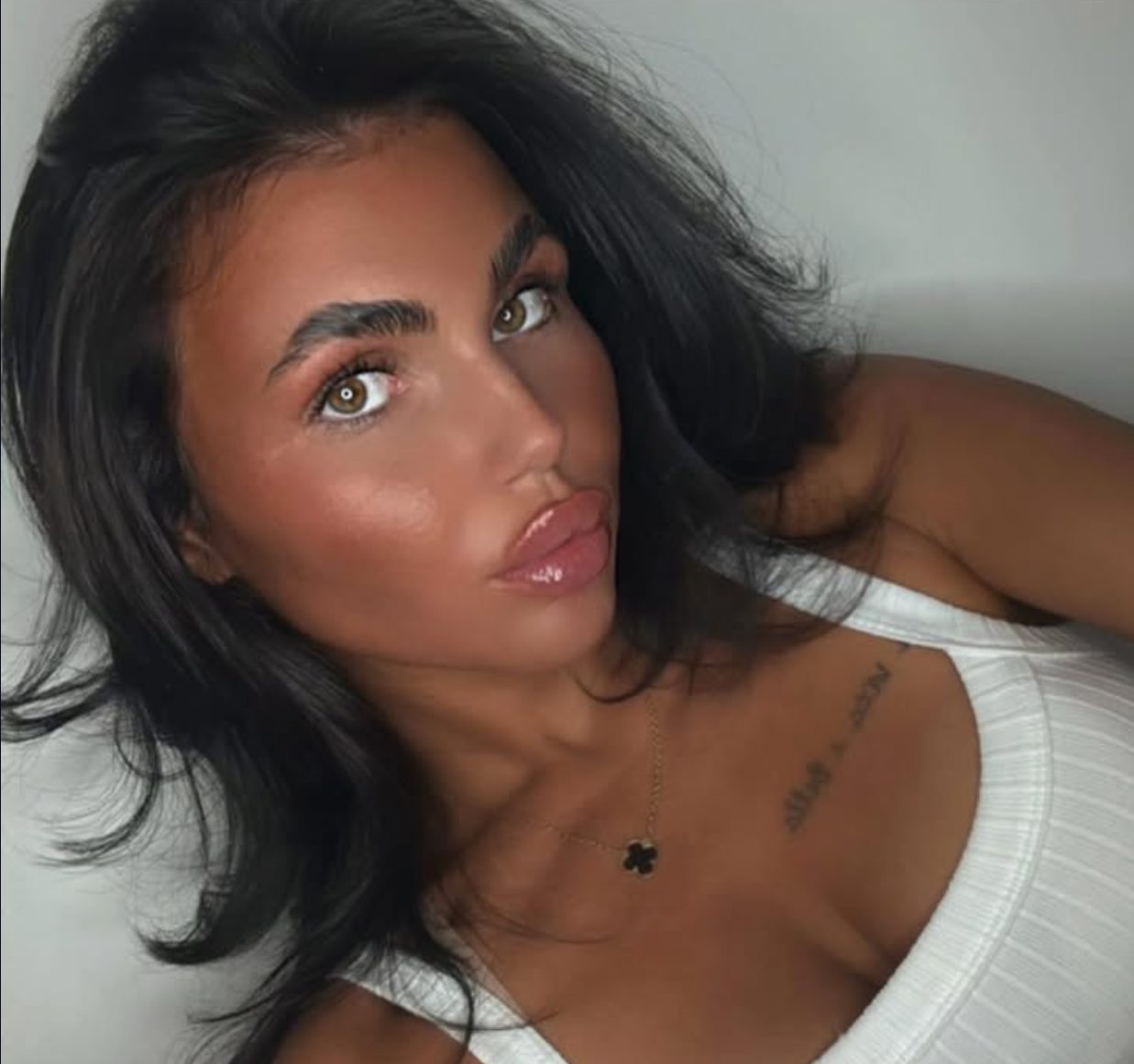 A woman with dark wavy hair, tan skin, and brown eyes, wearing a white top and a black four-leaf clover necklace, looking at the camera. RUSSIAN LIP FILLER