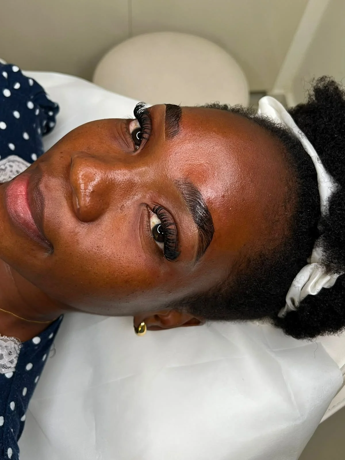 Close-up of a woman with dark skin lying on a bed, showing her face with freshly done eyelash extensions, dark eyebrows, and a white headband in her curly black hair.