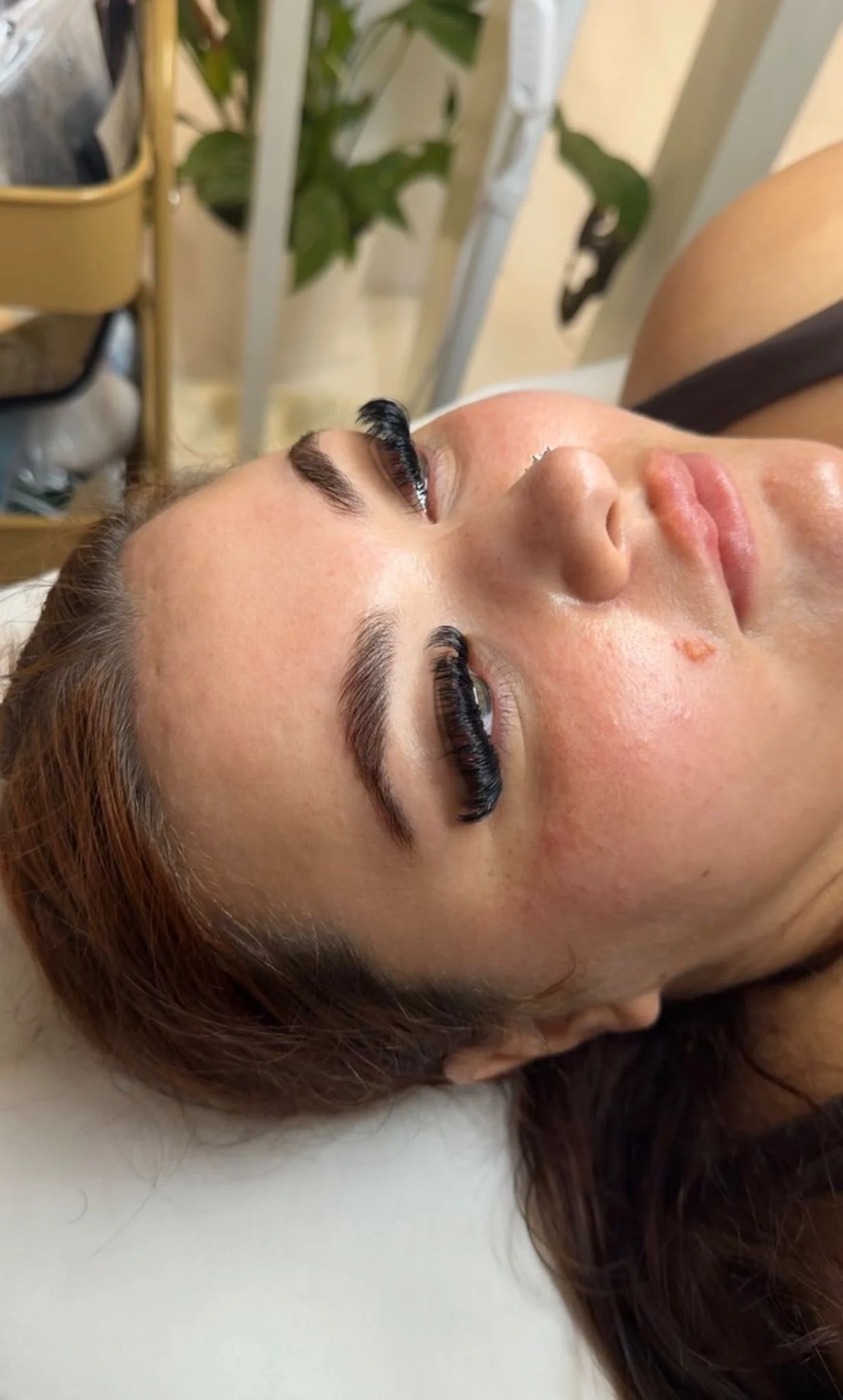 Close-up of a woman lying down with long hair, wearing eyelash extensions, with a nose piercing, in a beauty salon setting.