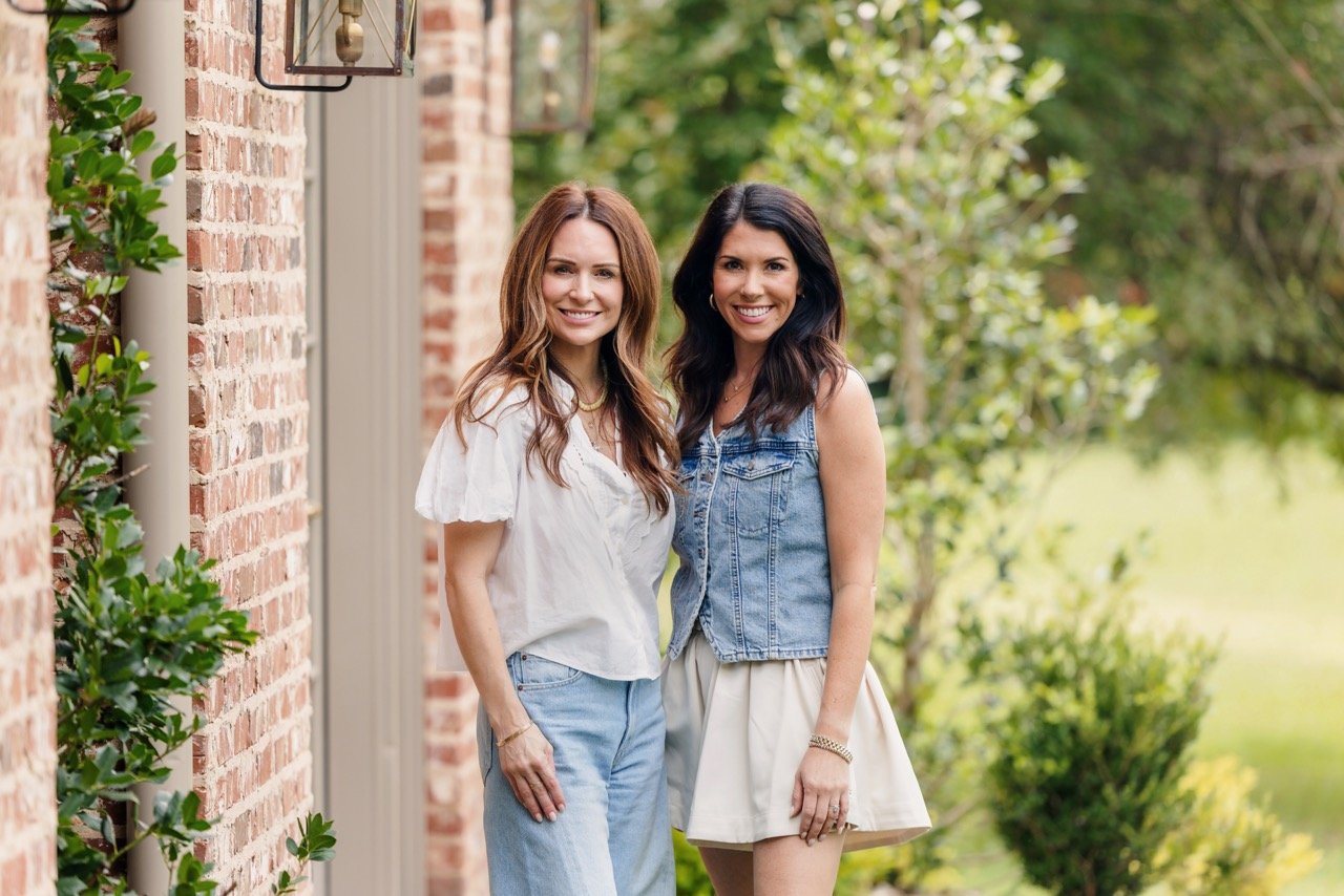 Two women standing outdoors near a brick wall, smiling, with greenery in the background.
