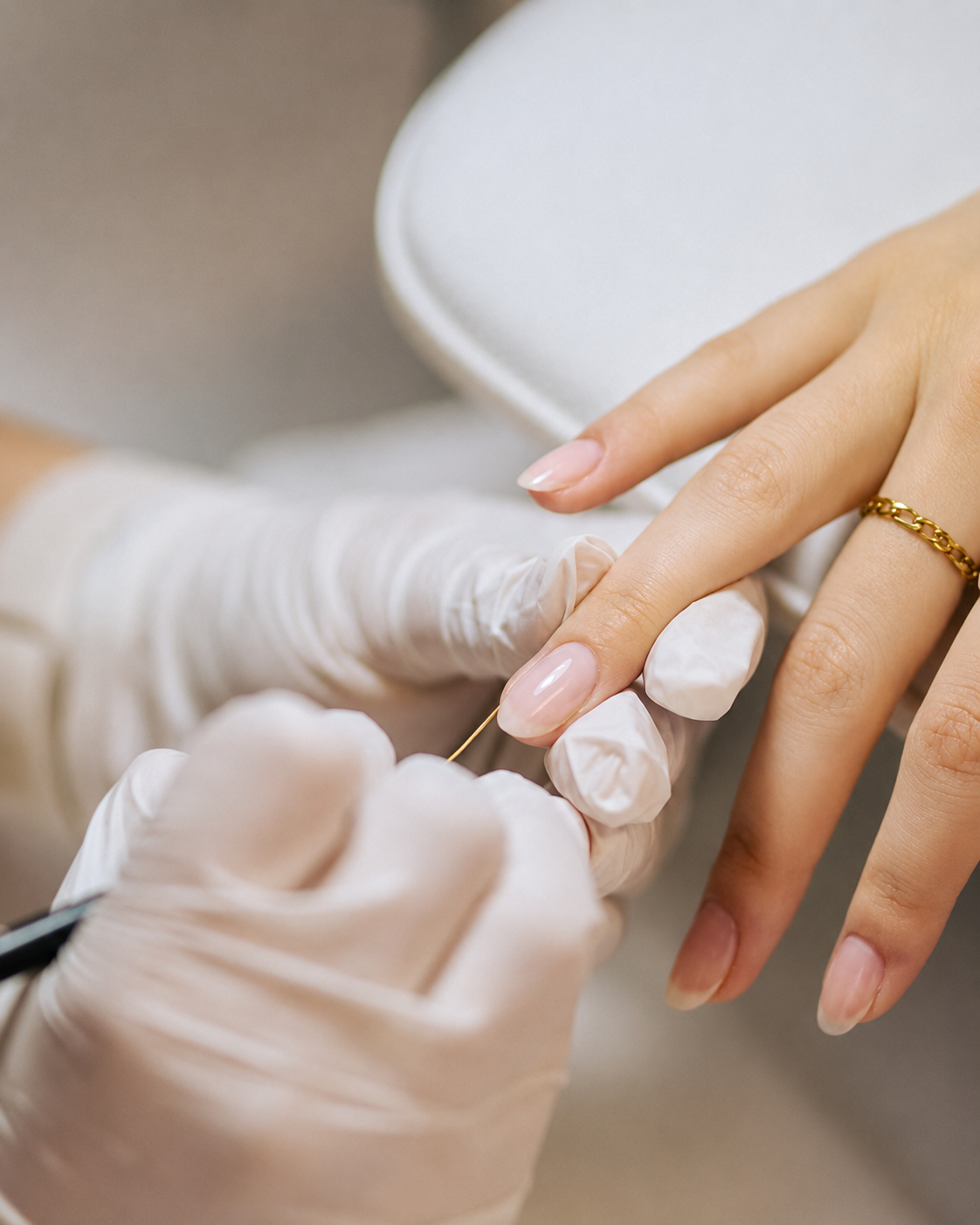 A person is receiving a manicure, with a nail technician applying polish or treatment to the client's fingernails. The client has a gold ring on their ring finger and is wearing a white top.