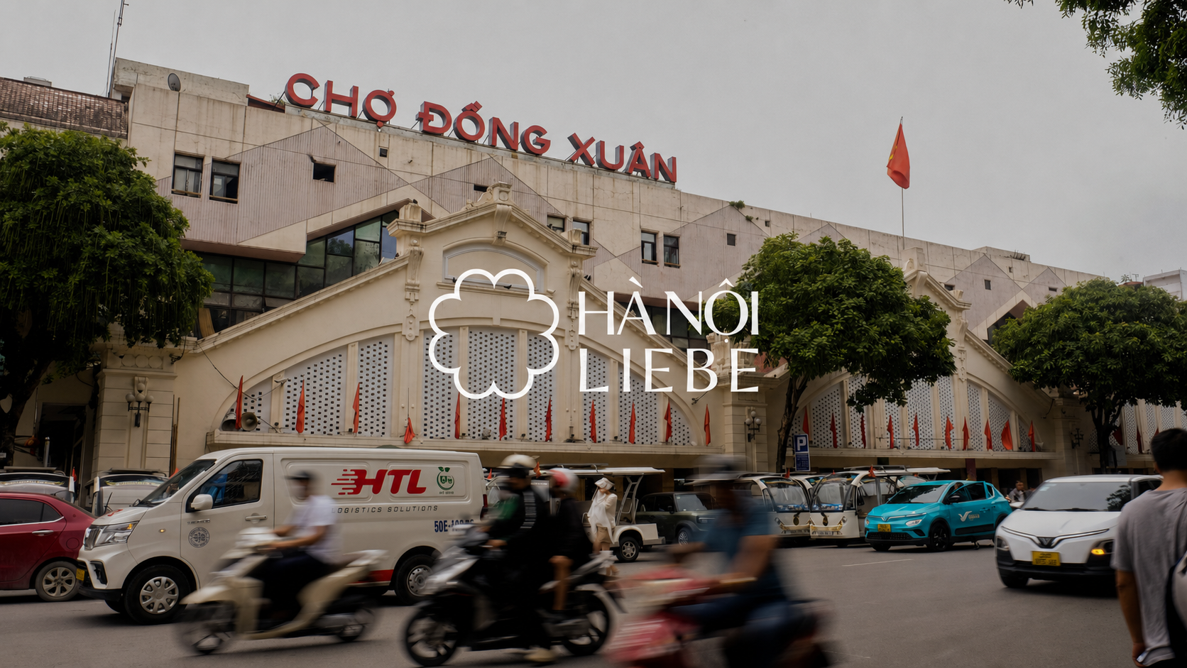 Street view of Chợ Đồng Xuân market in Hanoi, Vietnam, with cars and motorbikes on the road, trees, and a cloudy sky.