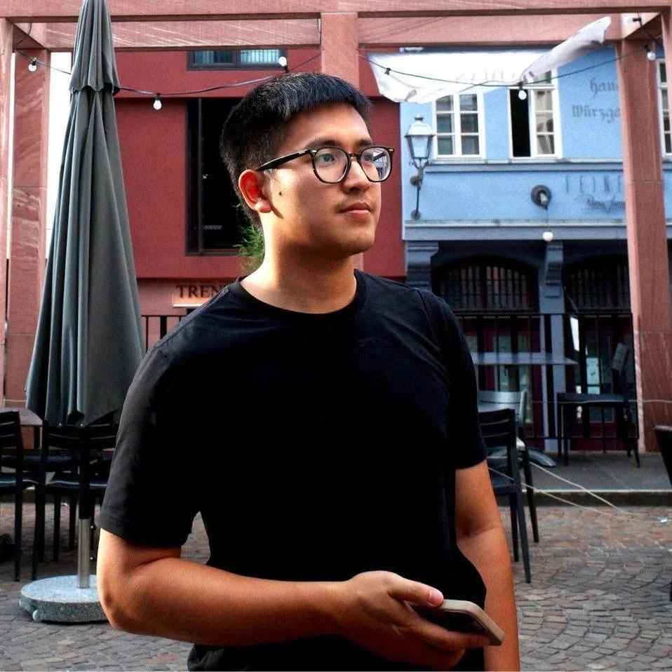 A young man wearing glasses and a black t-shirt is standing outdoors in a courtyard with brick pavement, holding his phone. Behind him are colorful buildings, outdoor tables, and string lights.