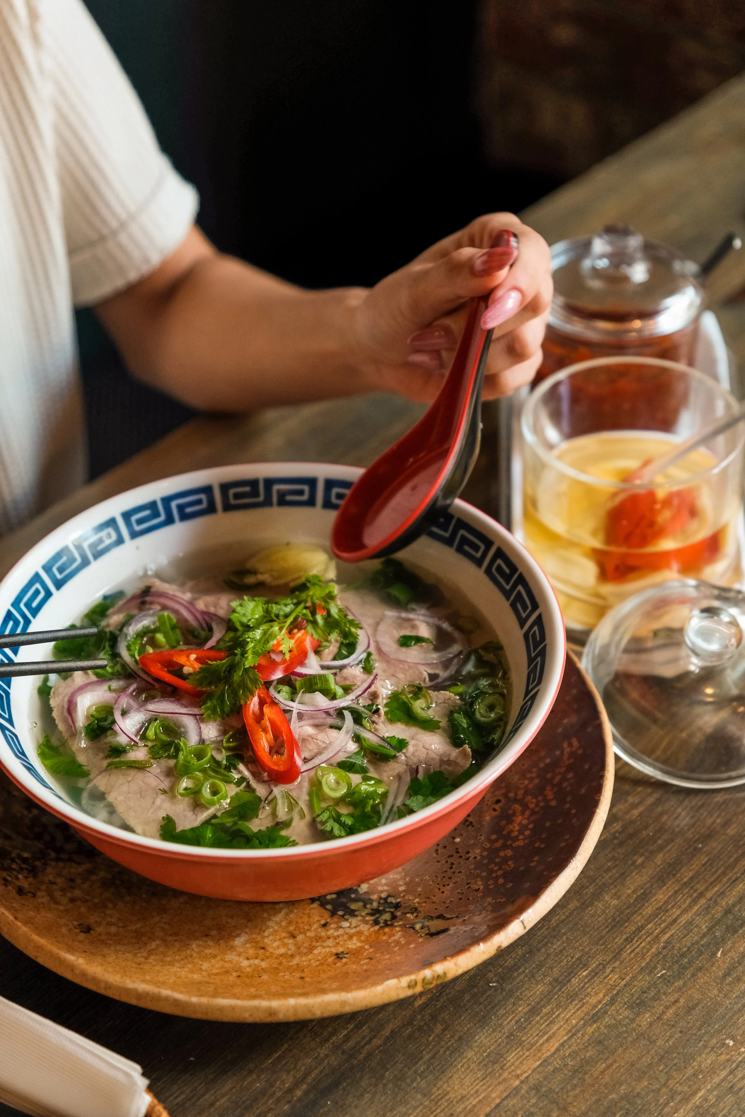 A bowl of Vietnamese pho topped with cilantro, sliced red chili peppers, and green onions, on a rustic wooden table, with condiments and drinks in the background.