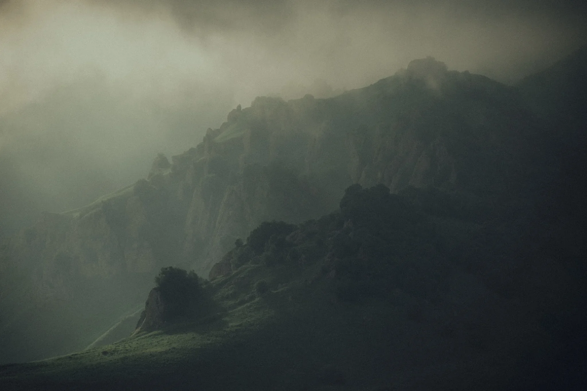 A mountain range shrouded in fog or clouds, with rocky peaks and a winding path or road in the foreground.