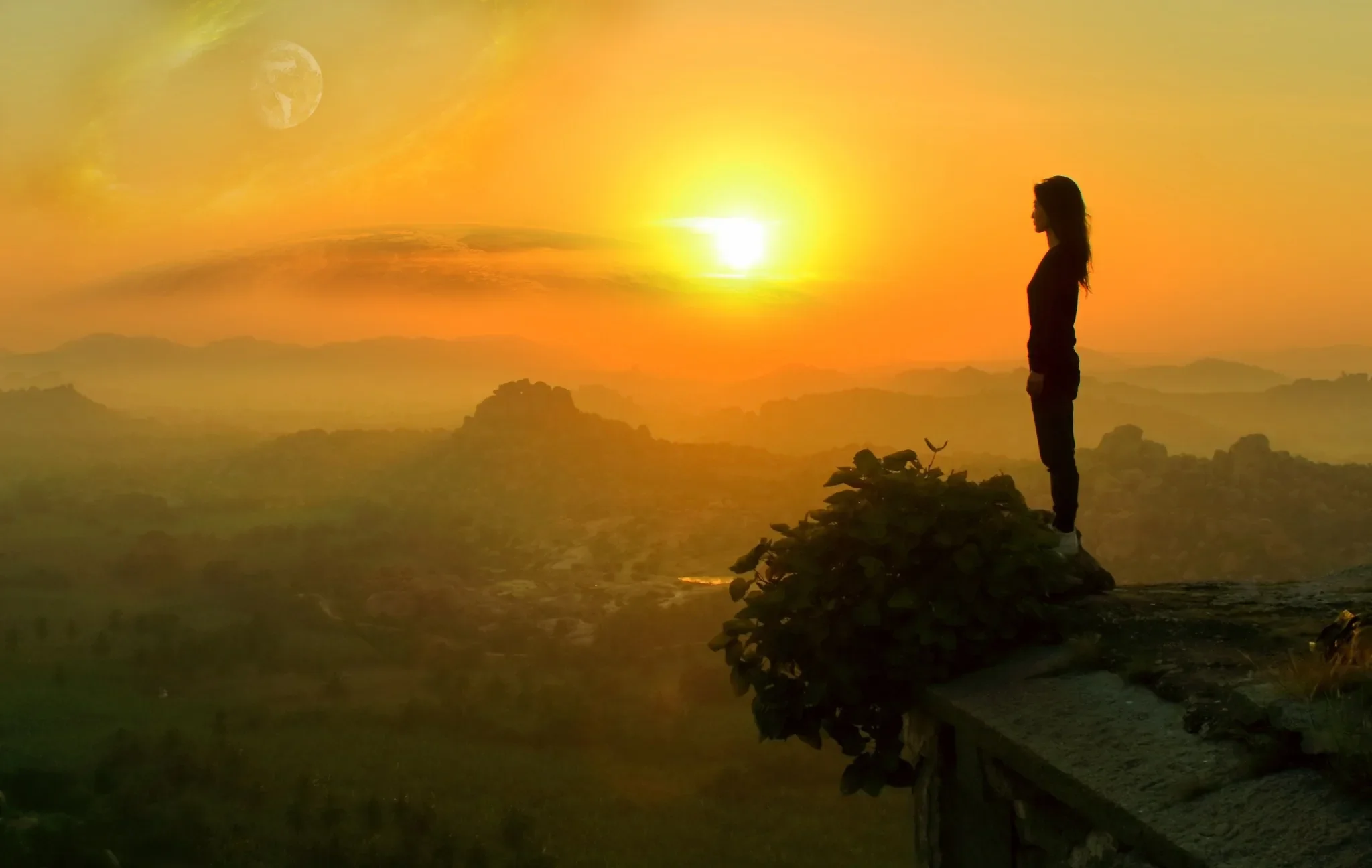 A person stands on a rocky ledge at sunset or sunrise, overlooking a landscape with mountains and a sky with a distant moon, illuminated by vibrant orange and yellow hues.